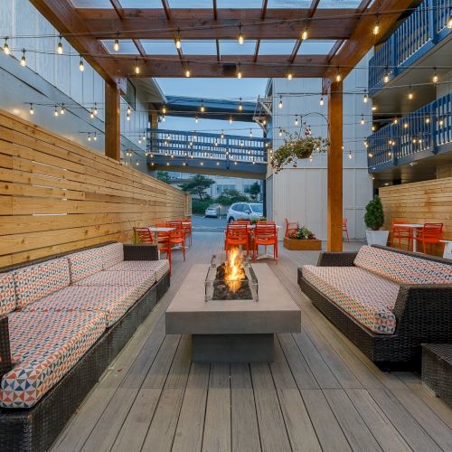 Outdoor patio with string lights, cozy cushioned benches, bright chairs, and a central fire pit under a wooden pergola.