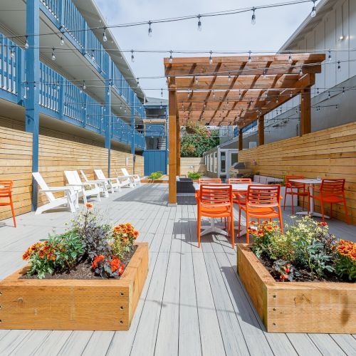 Sunlit outdoor patio with wooden planters, string lights, and orange chairs beside blue and white buildings.