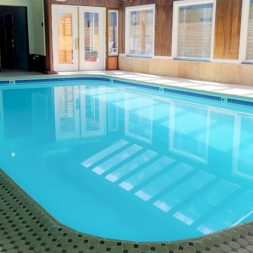An indoor swimming pool with blue water, tiled steps, a ladder, and doors and windows along the wall.