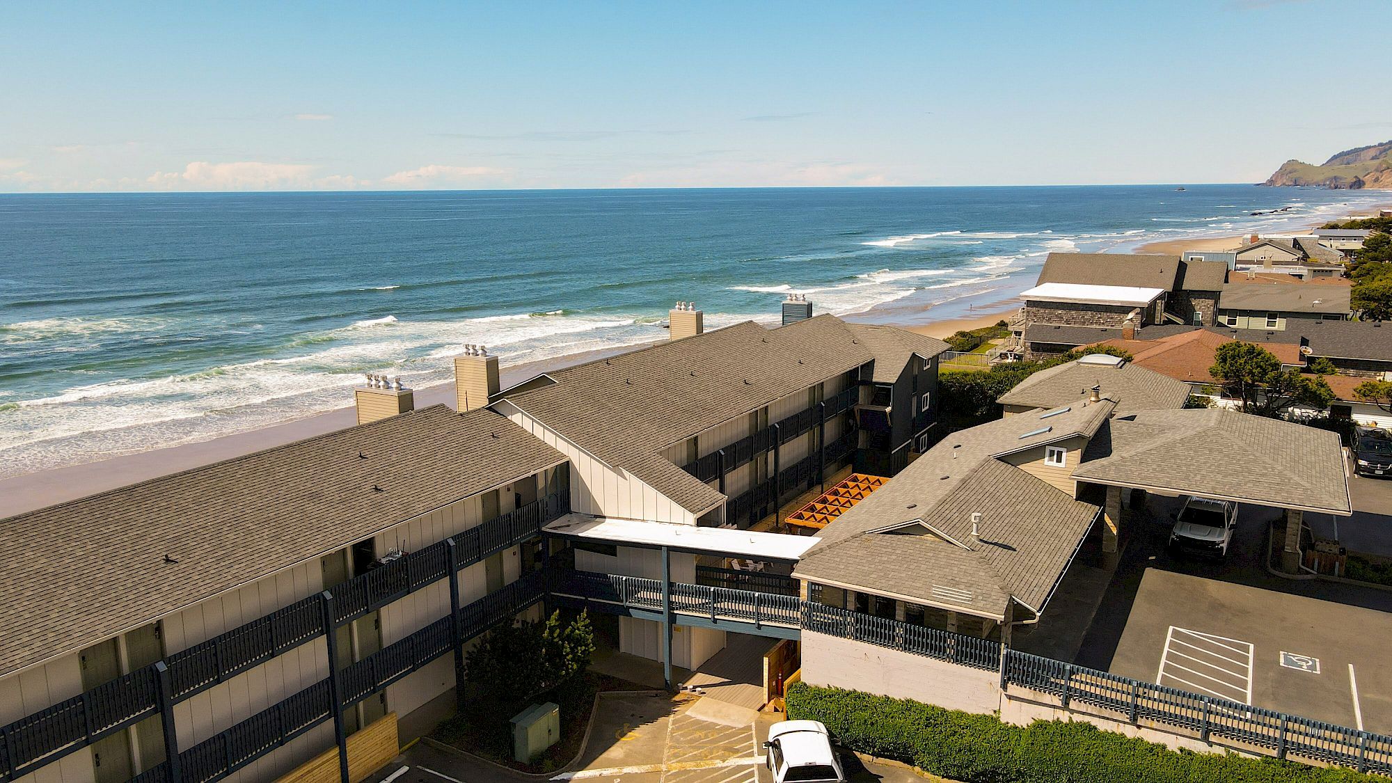 An aerial view of a beachfront motel with multiple buildings, ocean waves, and distant cliffs under a blue sky.