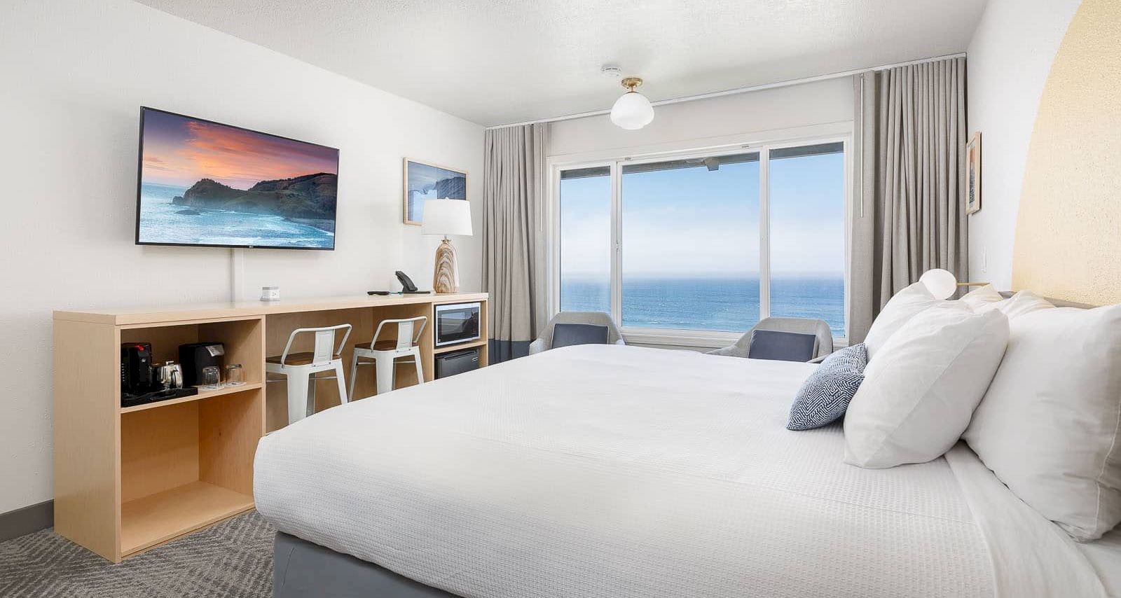 Bright hotel-style bedroom with a white bed, desk coffee setup, wall TV, and large windows showing the ocean view.